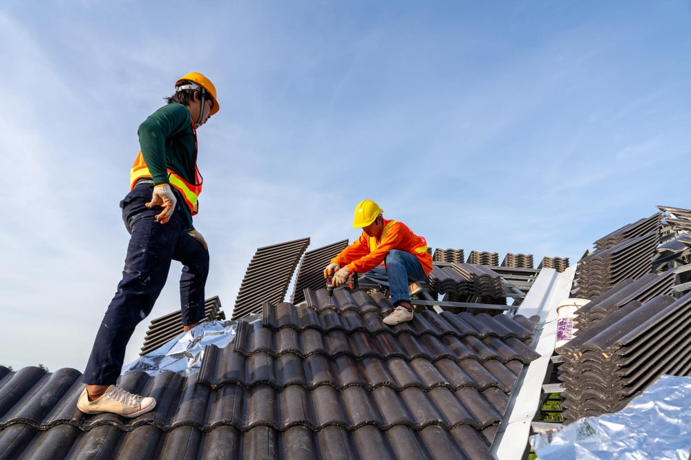 Two professional roofers in high-visibility safety gear and hard hats installing dark grey tiles on a sloped roof using power tools.